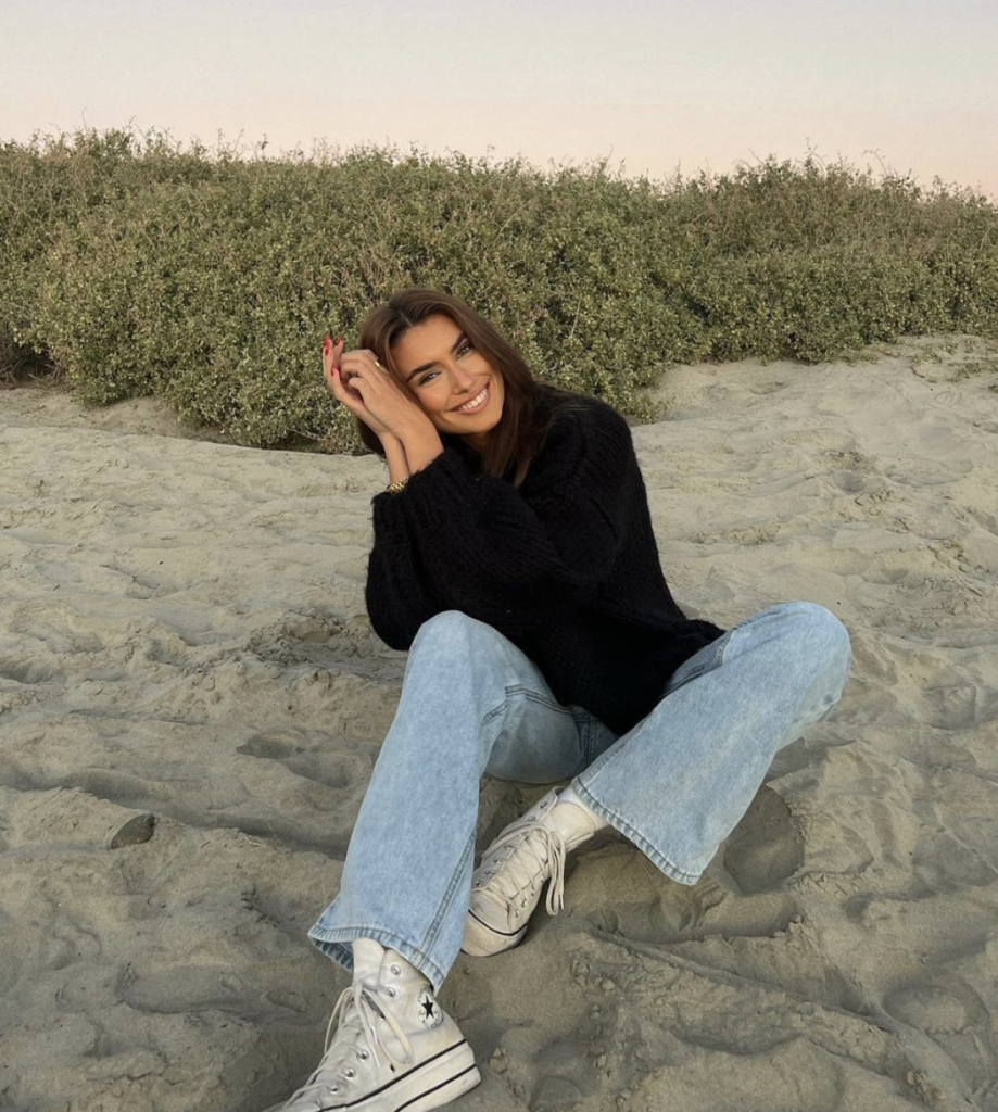 Sitting on the beach in a cozy black sweater, light-wash jeans, and classic high-top sneakers.